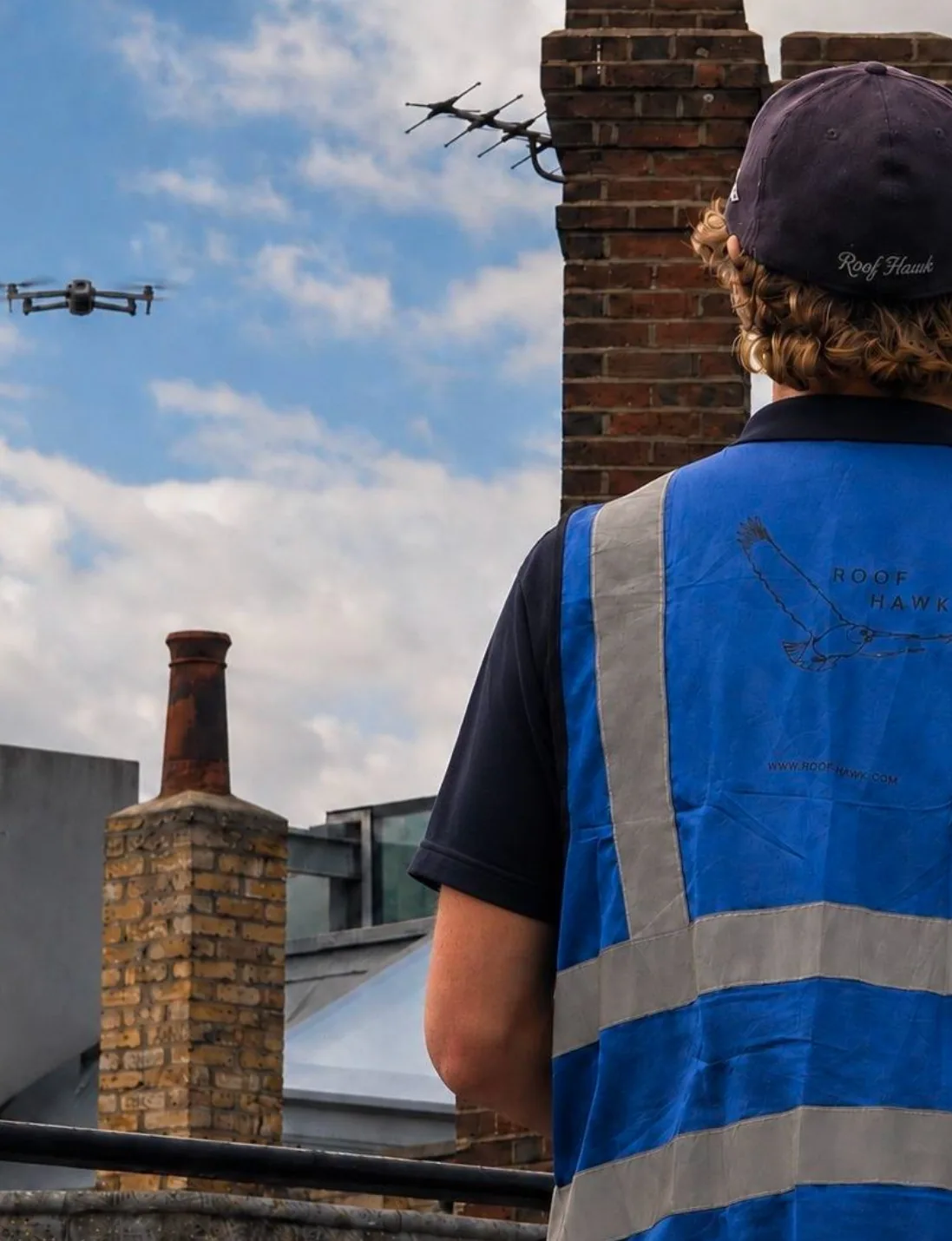 A Roof Hawk pilot in a branded cap and hi-vis vest watches the drone in flight beside brick chimney stacks on a London roof